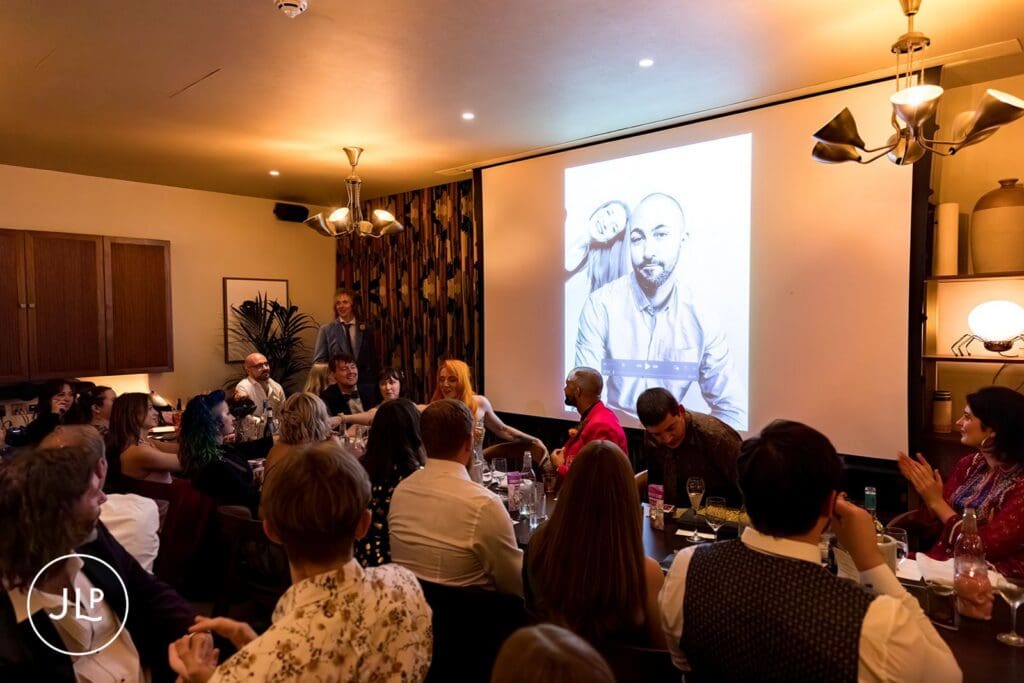 Wedding guests watching a slideshow presentation featuring a black and white portrait of the groom projected on a large screen.