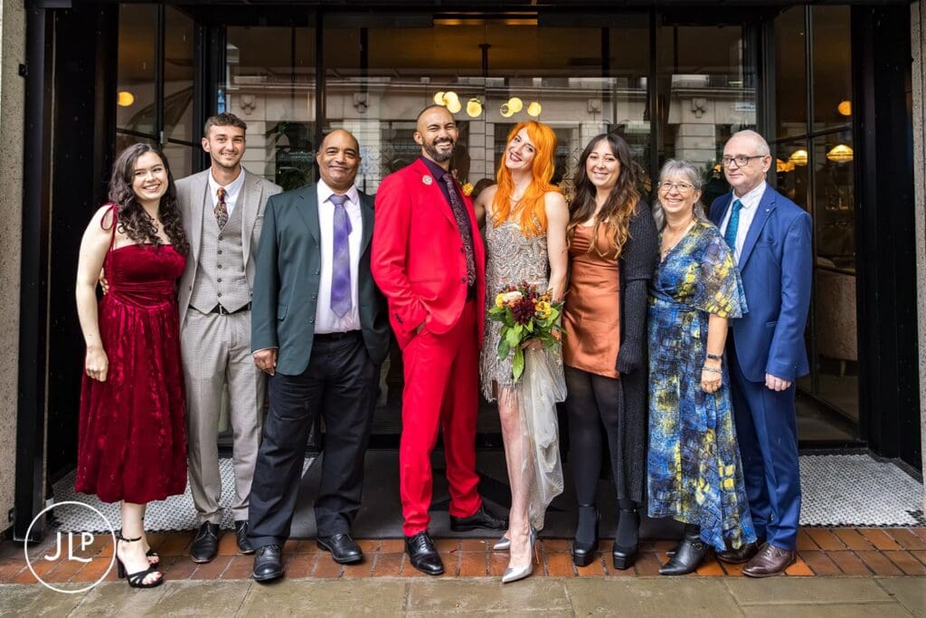 A formal group portrait of the newlyweds with their families standing outside the glass entrance of a London hotel.