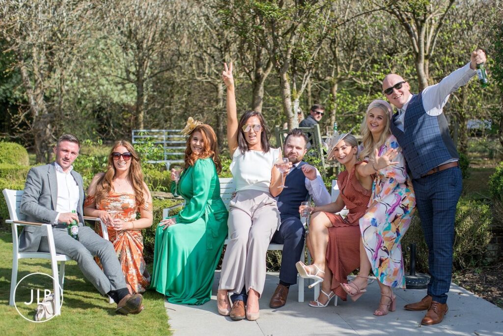 A group of wedding guests laughing and enjoying drinks on a garden bench during a sunny afternoon reception at Langshott Manor.