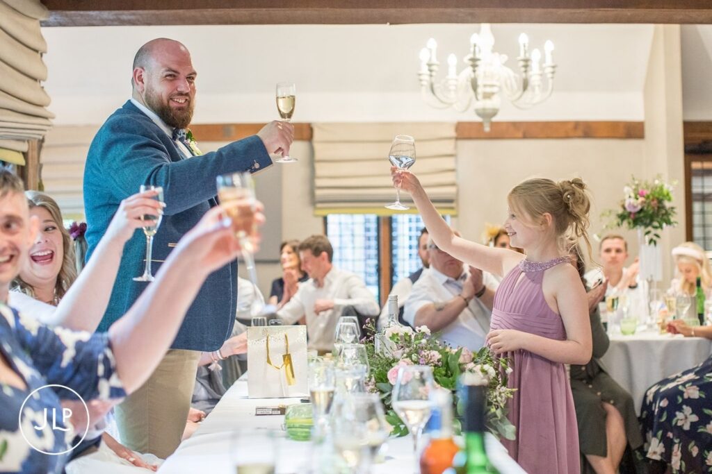 A young bridesmaid raising a glass for a toast with the groom during a wedding breakfast at Langshott Manor in Surrey.