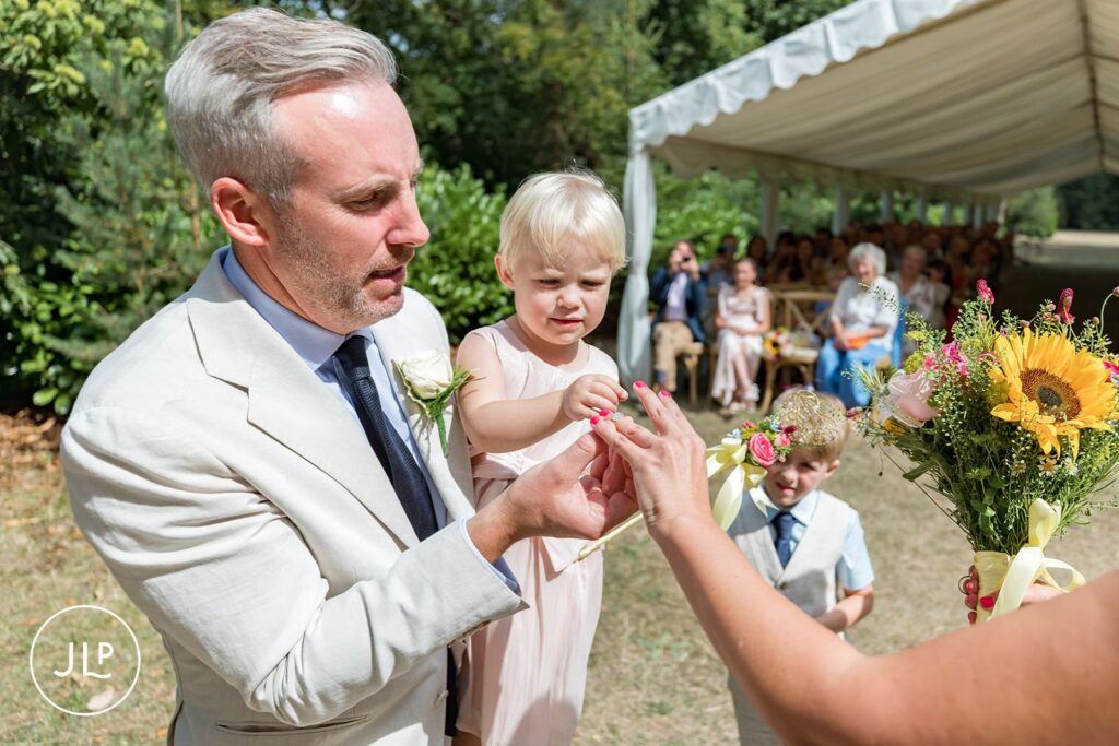 Candid wedding photography of a flower girl helping the groom place a ring on the bride's finger.