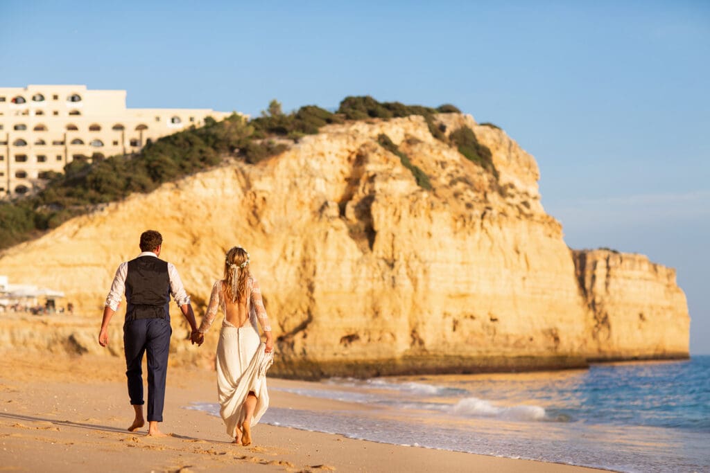 Newlyweds walking barefoot on a beach near Carvoeiro, shot by an Algarve beach wedding photographer.