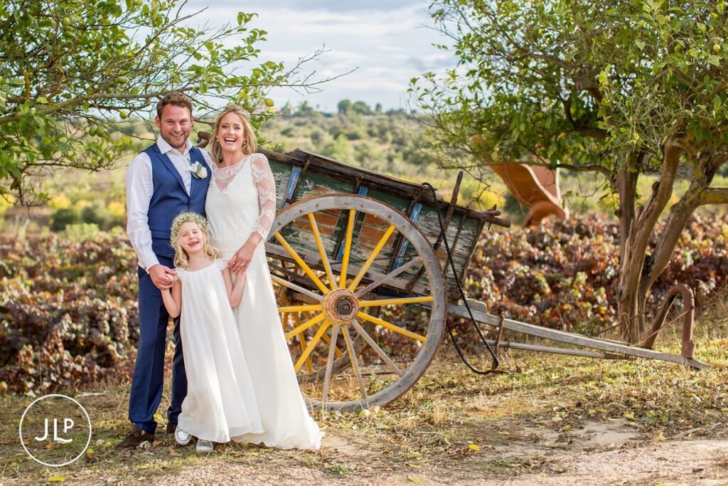 Ali, Matty, and a young girl posing by a rustic wooden cart during an Algarve vineyard wedding.