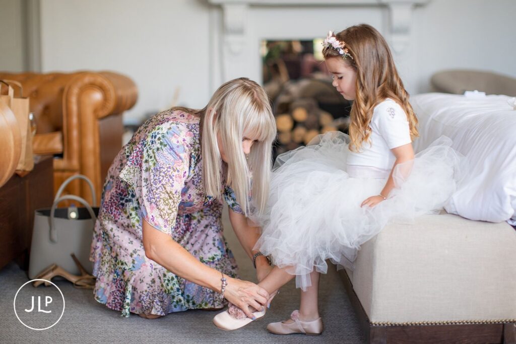 A woman in a floral dress helps a young flower girl put on her shoes for a luxury Gloucestershire wedding.
