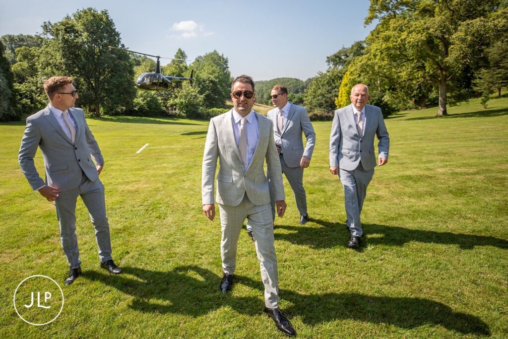 Five groomsmen in matching grey suits walk across the lawn, with a helicopter in the background.