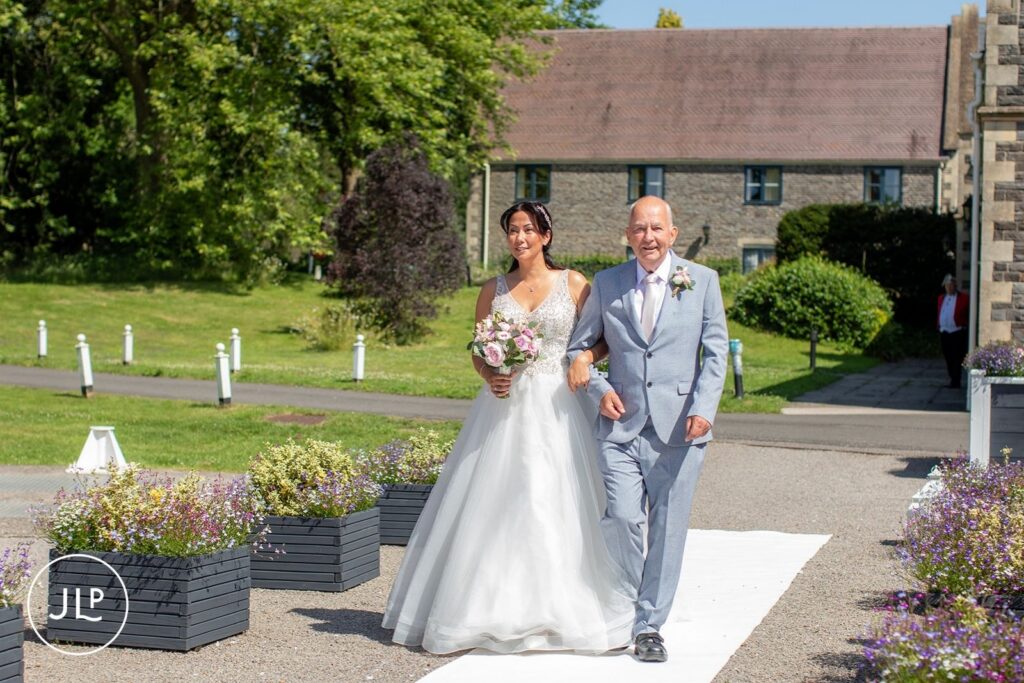 The bride carries a pink rose bouquet as she is walked down the outdoor aisle by her father.