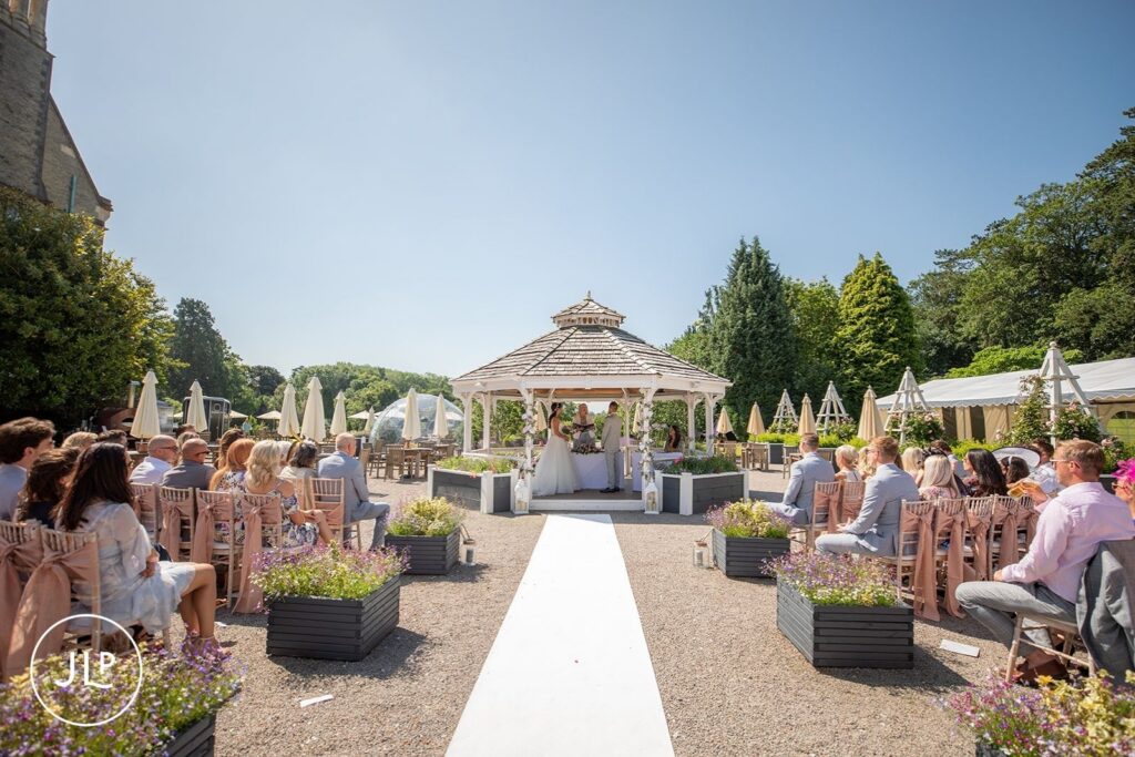 A wide-angle view showing the white aisle runner, wooden gazebo, and seated guests during an outdoor ceremony.