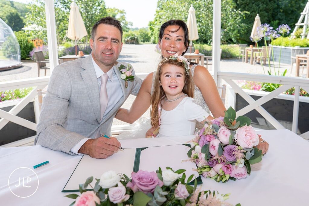 Mel and Ben smile while seated with a flower girl as they sign their wedding certificates.