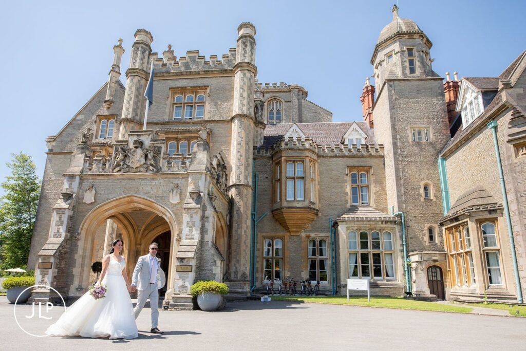 A wide shot of the couple walking past the grand stone entrance of the luxury Gloucestershire wedding venue.