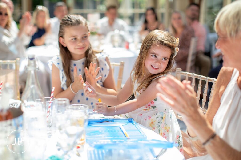 Two young flower girls smile and clap while sitting at a decorated table during a natural wedding photography session.