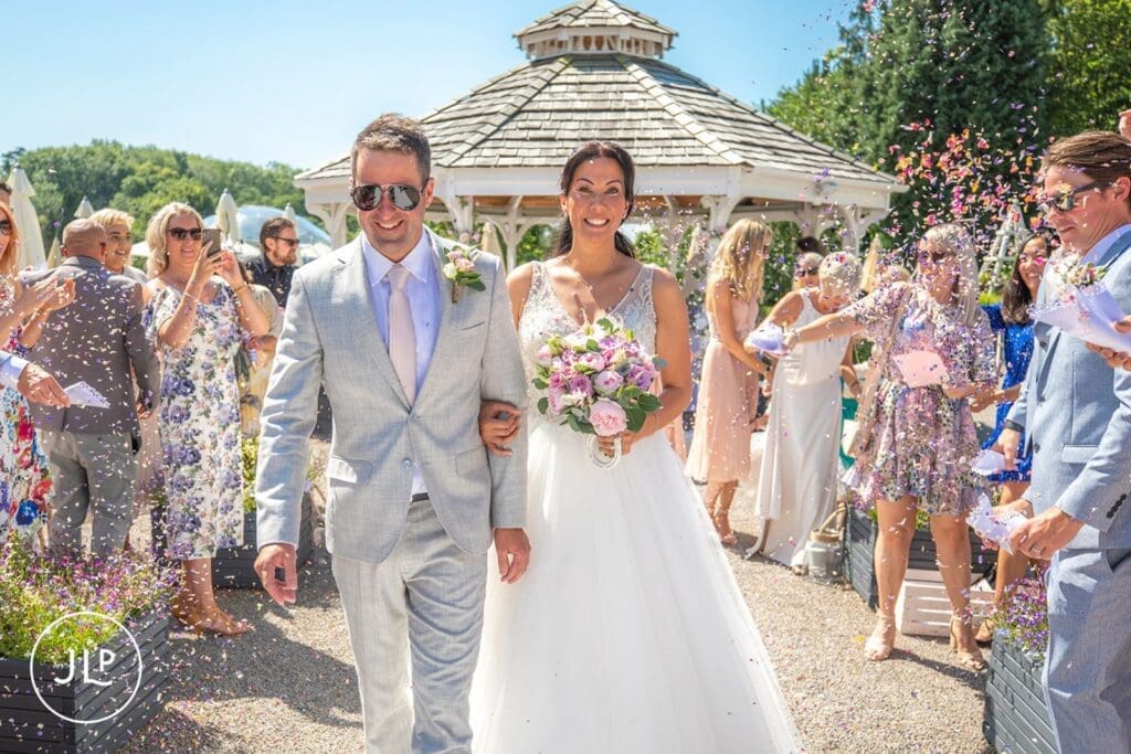 Bride and groom smiling through a confetti tunnel by an outdoor wedding photographer UK.