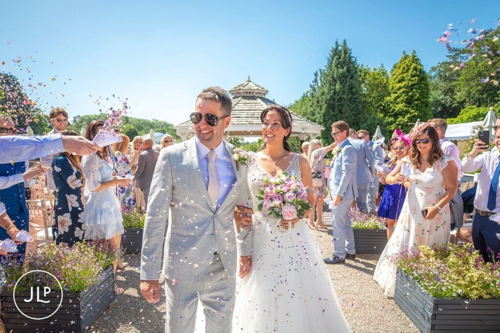 Smiling bride and groom showered in colourful confetti by guests under a bright summer sky.