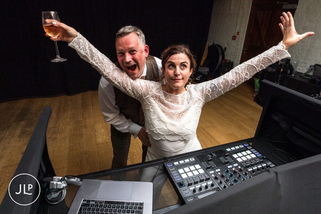 Bride in a white jumpsuit and groom posing behind the DJ decks during a fun autumn wedding at Wasing Park.