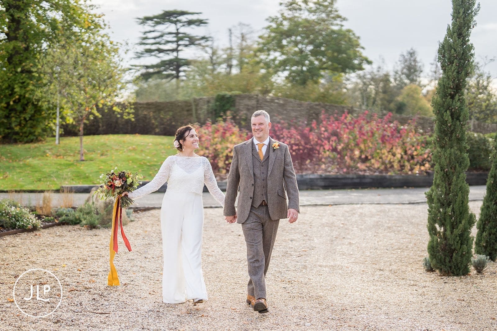 Newlyweds holding hands and smiling on a gravel path during their autumn marriage celebration.