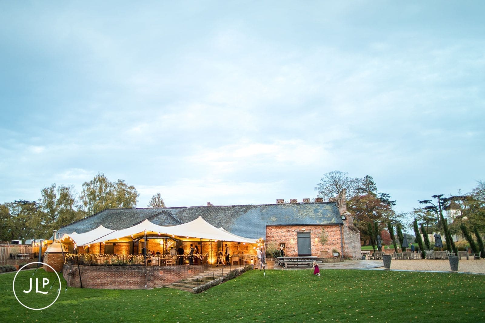 Exterior view of a large wedding marquee lit up with fairy lights on a sprawling green lawn at dusk.