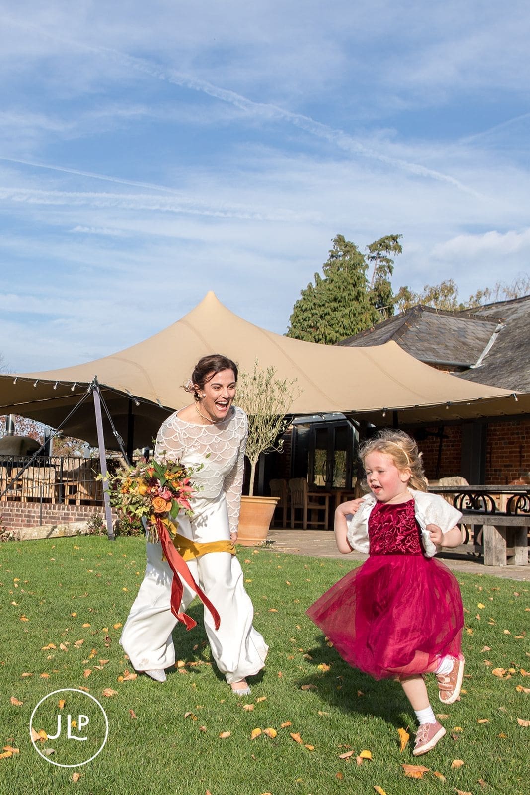 Smiling bride in a modern white jumpsuit chases a flower girl on the lawn at an autumn Wasing Park wedding.