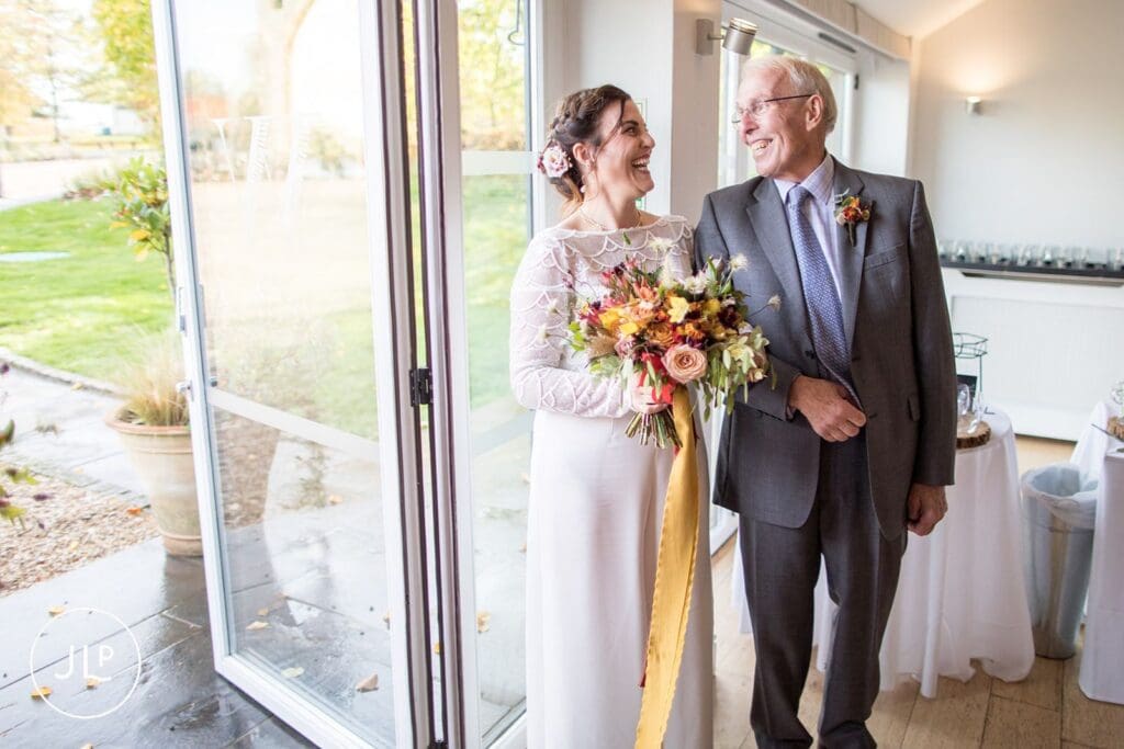 Candid wedding photography of a bride in a modern white jumpsuit laughing with her father at Wasing Park.