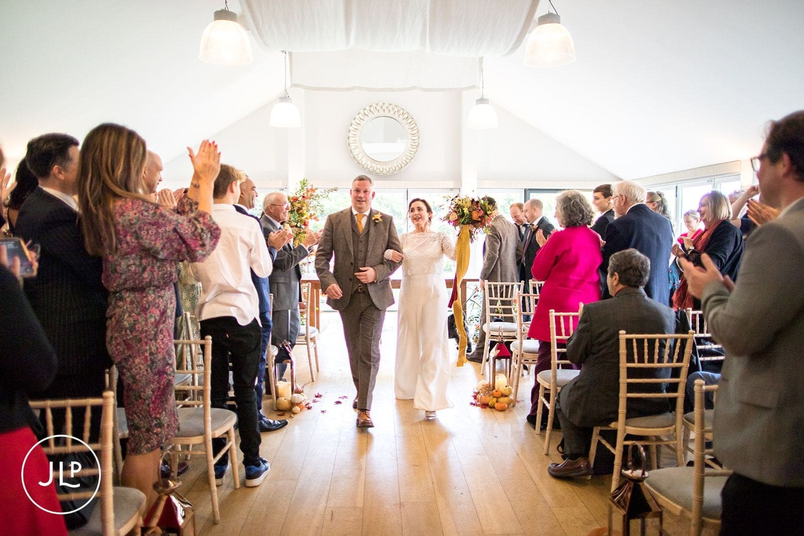 Newlyweds walking down the aisle together as guests applaud at a stylish indoor autumn wedding ceremony.