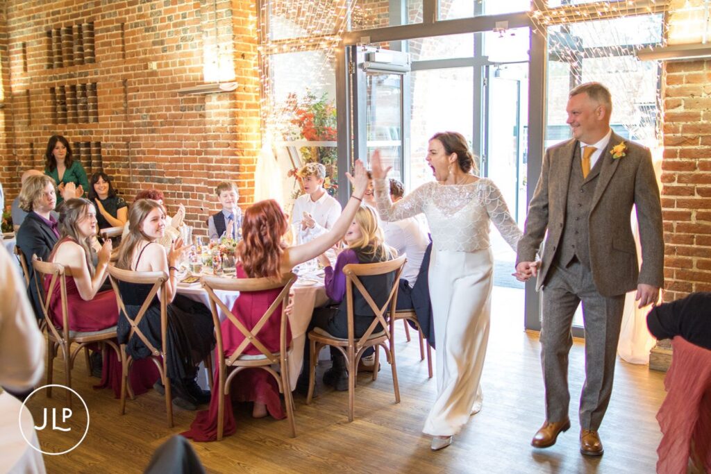 Bride in a stylish white jumpsuit high-fives guests during an entry, by a Wasing Park wedding photographer.