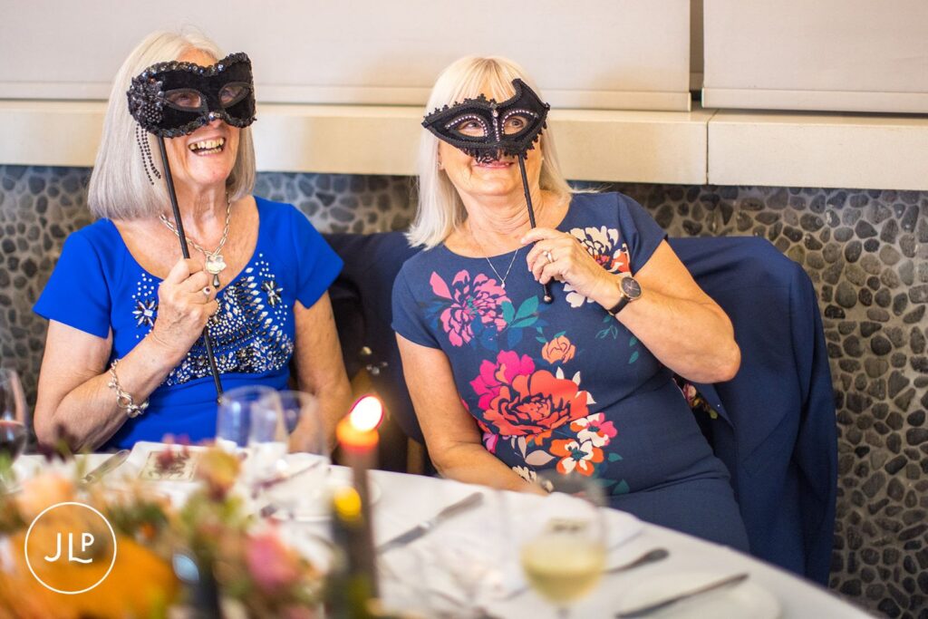Two wedding guests laughing while holding black masquerade masks at a relaxed wedding breakfast in Berkshire.