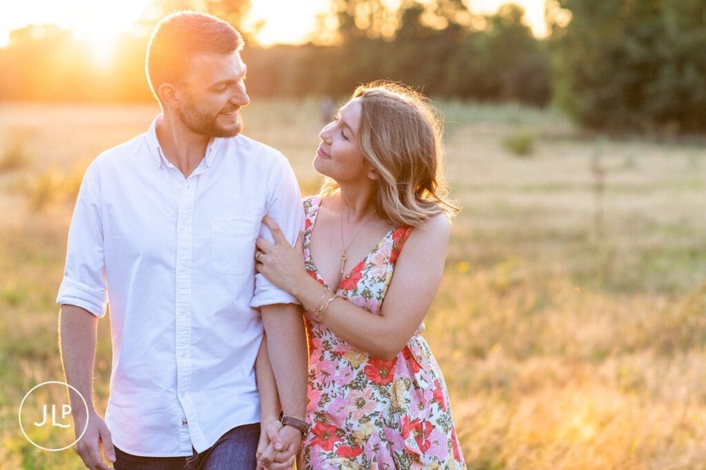 London engagement photographer captures a couple walking arm-in-arm through a golden sunset field.