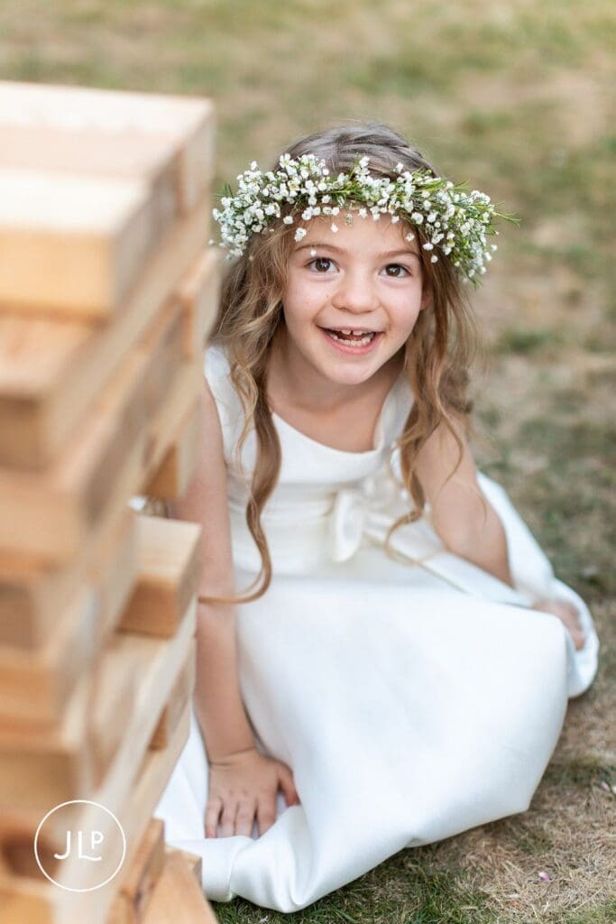 Smiling flower girl wearing a delicate floral crown next to giant Jenga at a Surrey wedding venue.