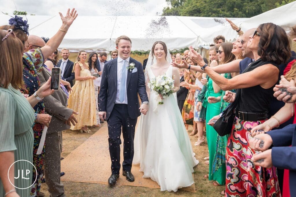 Candid wedding photography of a bride and groom walking through confetti at a Surrey marquee wedding.