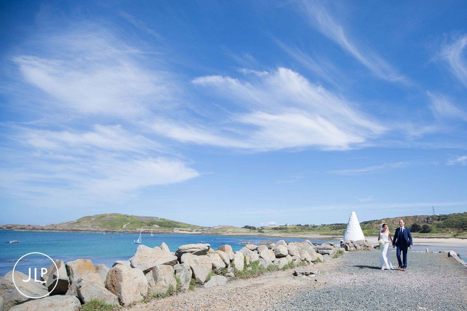 Wide shot of a bride and groom walking by the sea wall under blue skies, beautiful Alderney wedding photography.