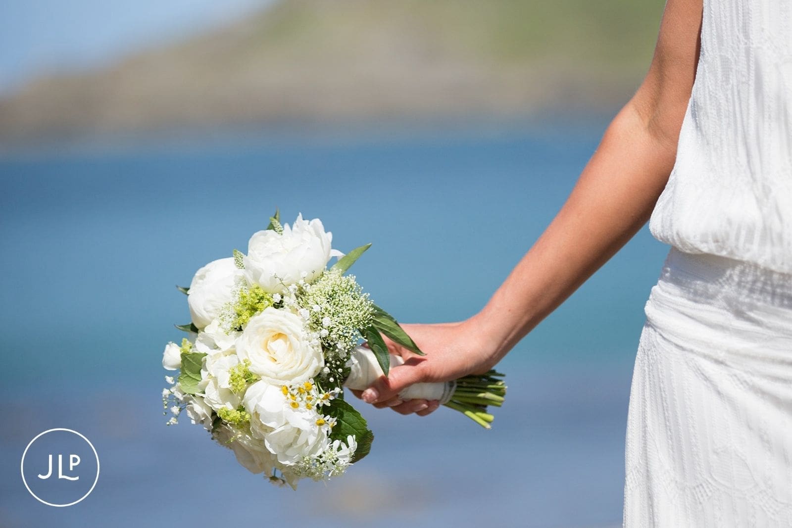 Bride gently holding her white wedding bouquet against a bright blue coastal ocean background in Alderney.