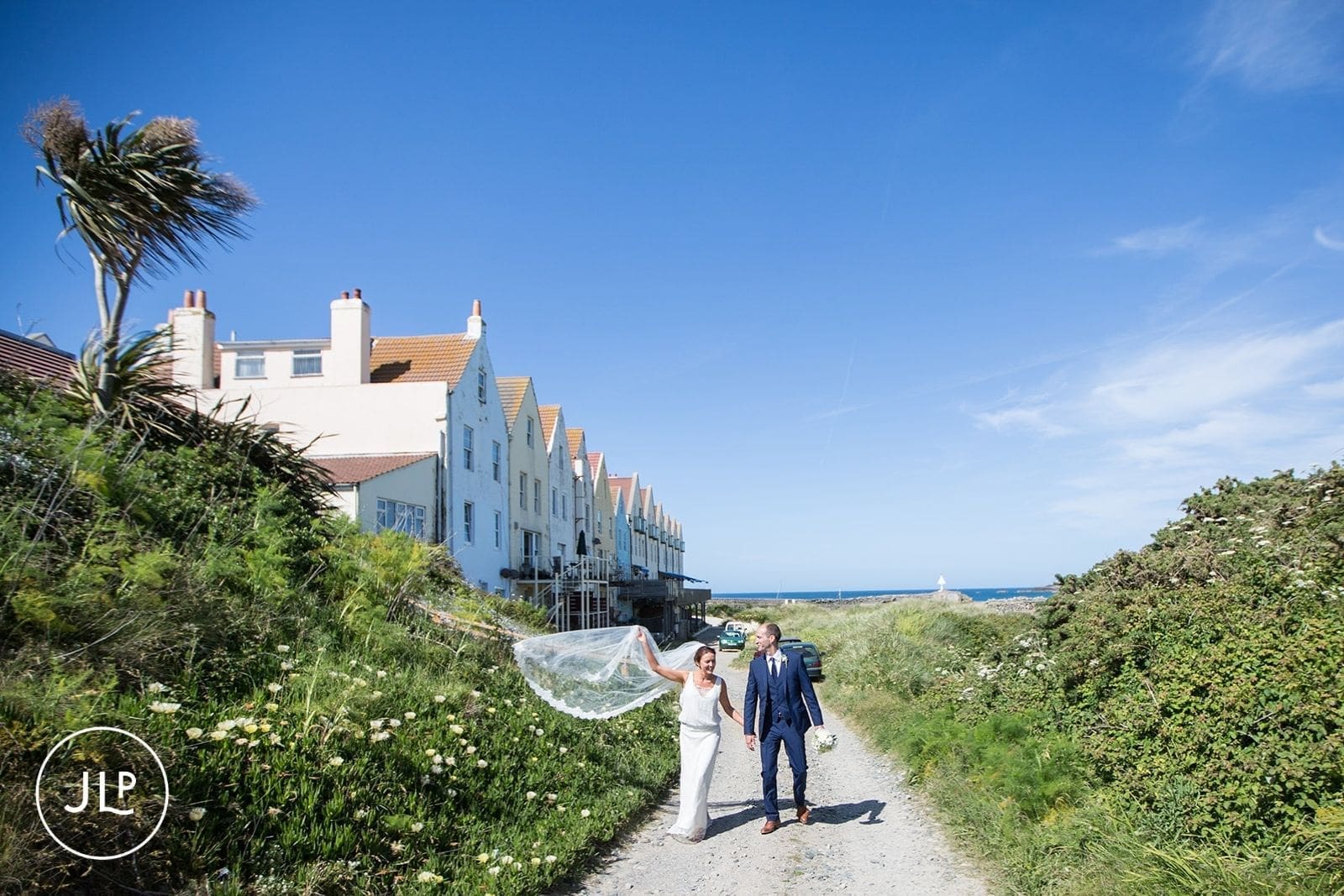 Newlyweds walking down a country road while the bride's veil catches the wind, getting married in Alderney.