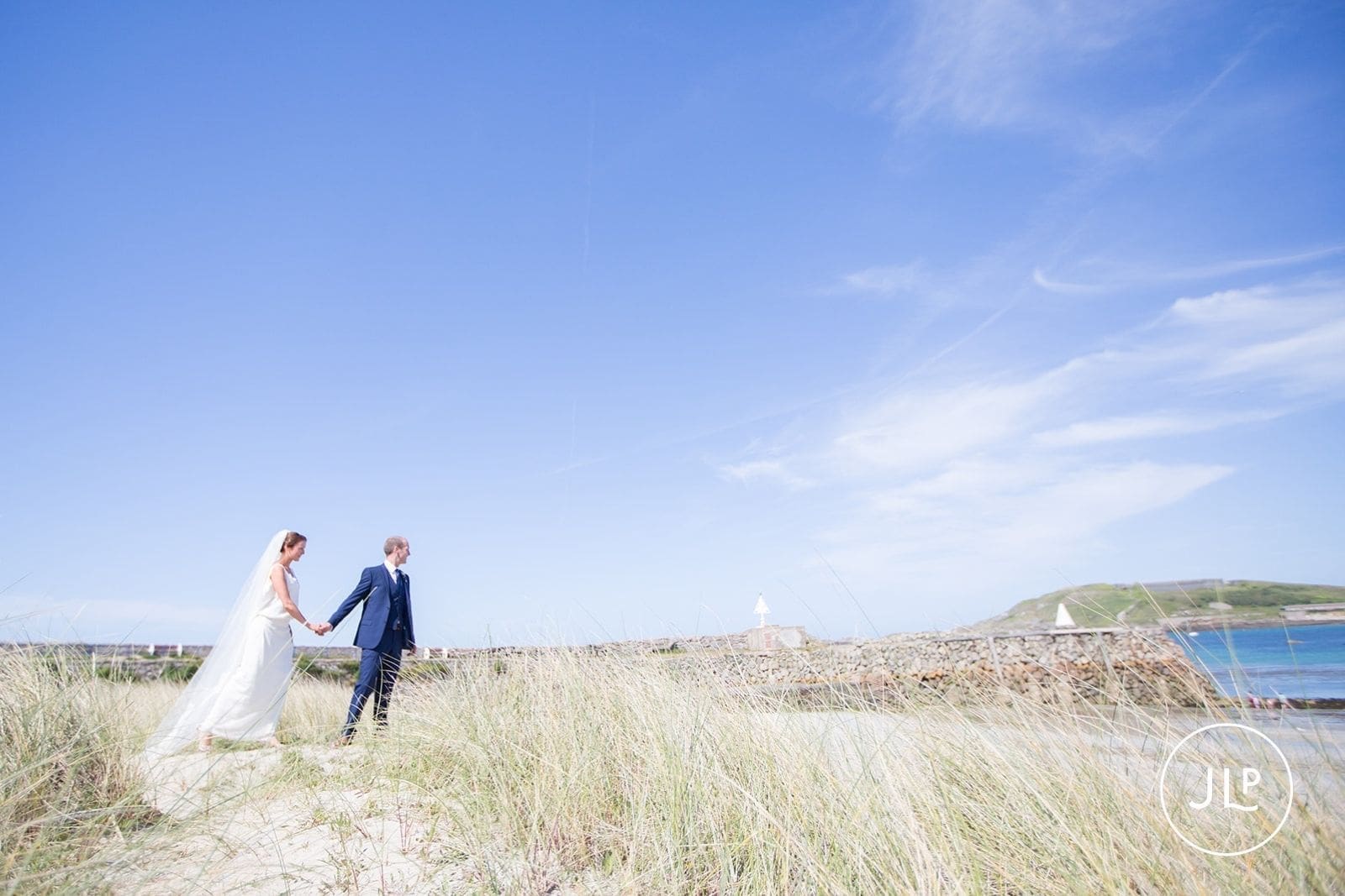 Romantic bridal photography of a couple holding hands and walking through grassy beach dunes in Alderney.