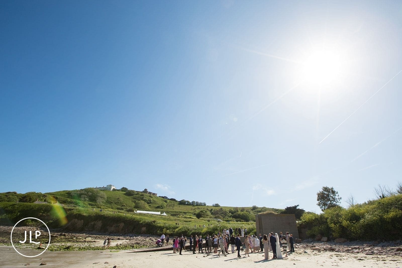 Sun-drenched documentary wedding photography of guests gathering on a beach below a rolling green hillside.