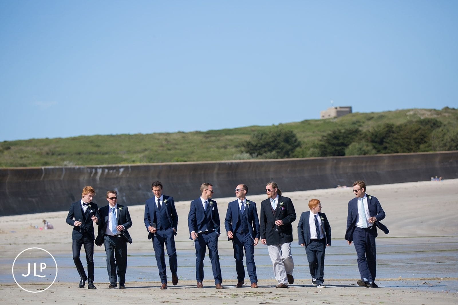 Groomsmen walking along a wide sandy beach together during a scenic destination wedding in Alderney.