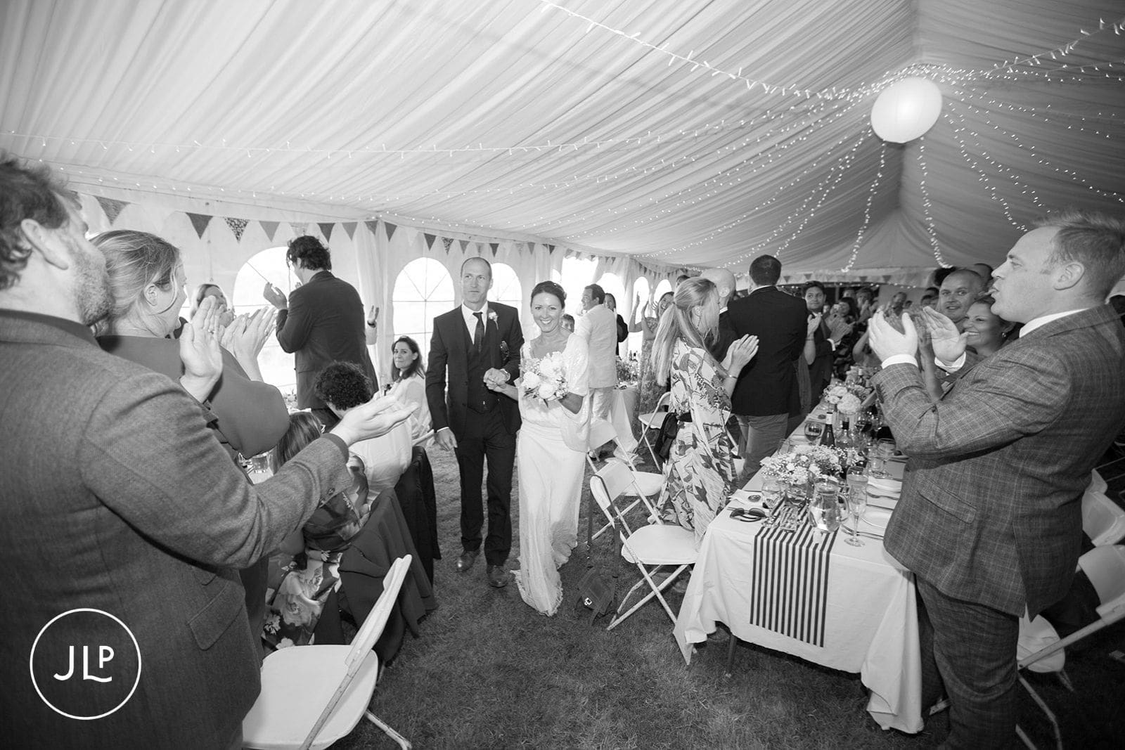 Black and white photo of newlyweds entering a marquee tent reception to cheering wedding guests.