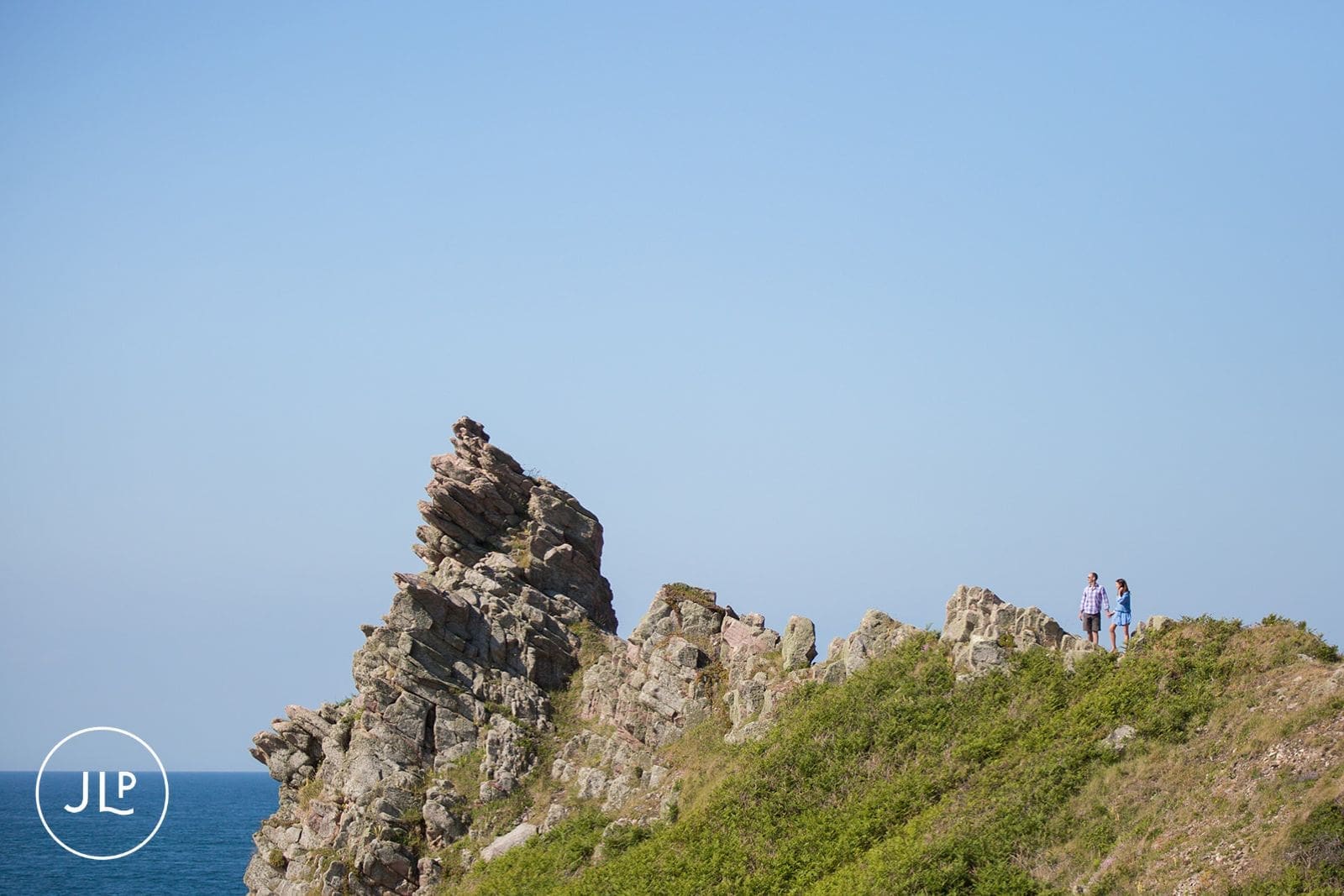 Distant shot of a couple standing on top of a dramatic rocky cliff by the blue sea.