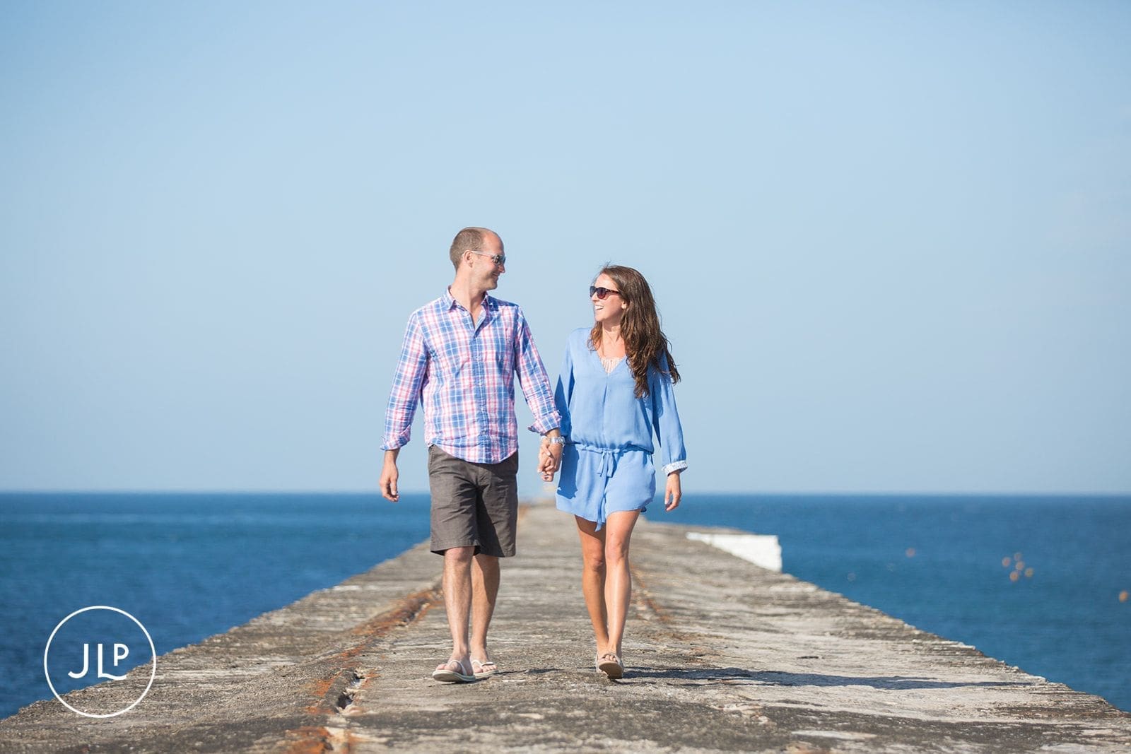 Couple holding hands and walking down a stone pier during a relaxed coastal engagement session.