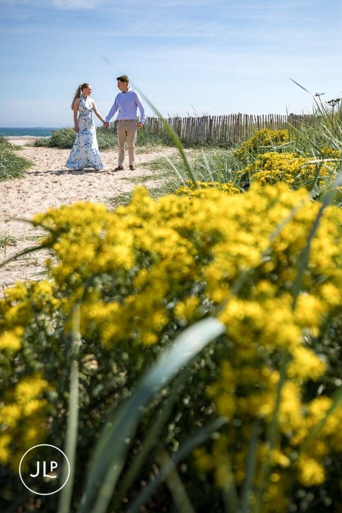 Romantic pre-wedding photos of a couple walking behind bright yellow beach flowers.