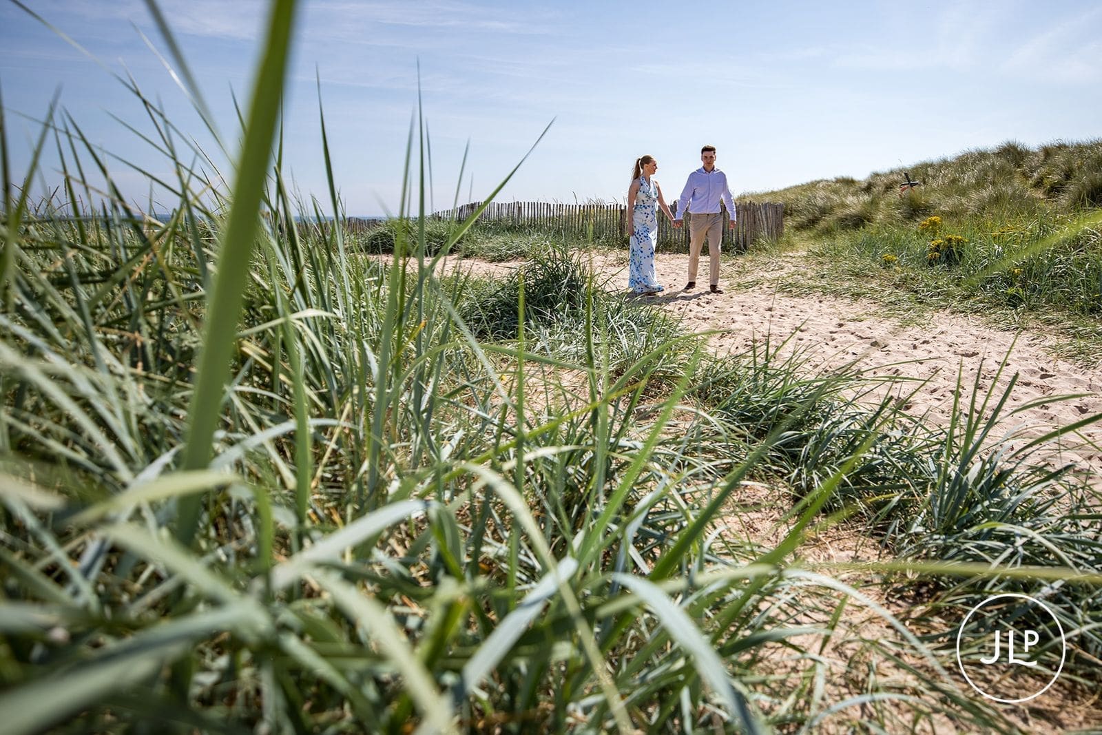 Seaside engagement photography of a couple walking together behind tall beach grass.