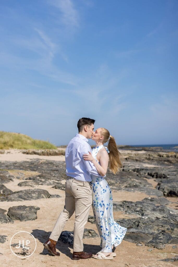Romantic beach engagement shoot of a couple kissing on a rocky shoreline under a bright blue sky.