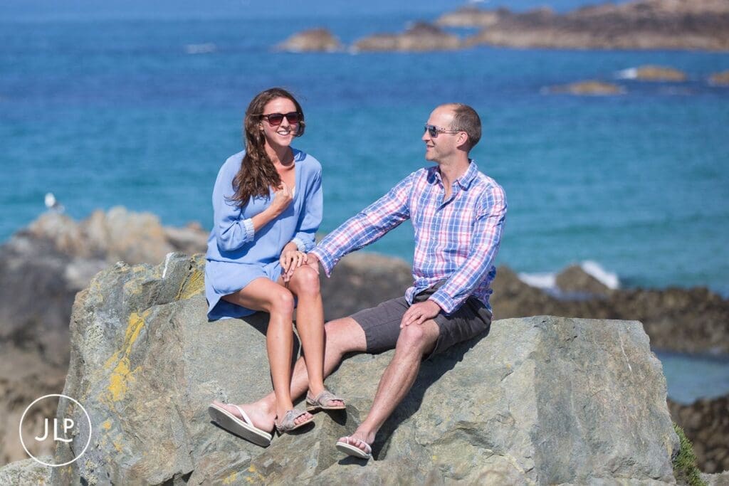 Happy couple sitting on coastal rocks smiling during their island seaside portrait session.