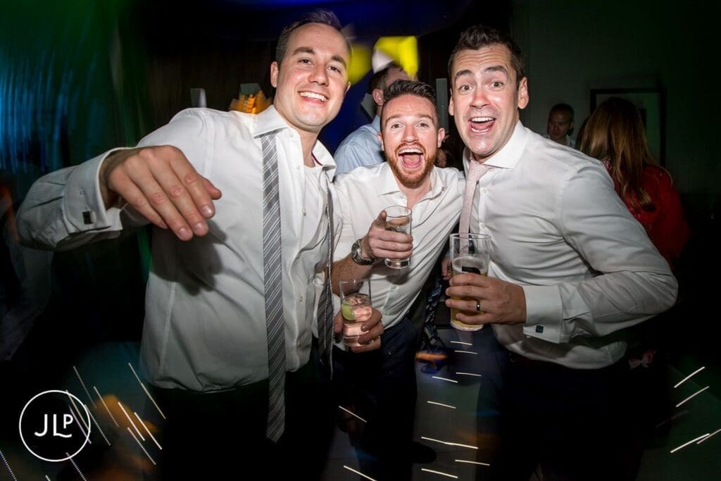 Energetic groomsmen dancing and celebrating with drinks on a lively evening wedding reception dance floor.