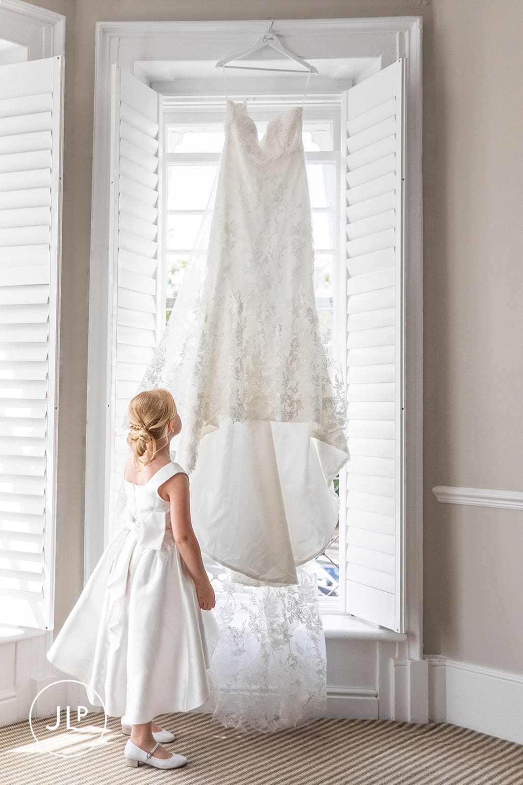 A young flower girl looking up at a beautiful bridal gown hanging by a window before the wedding ceremony.
